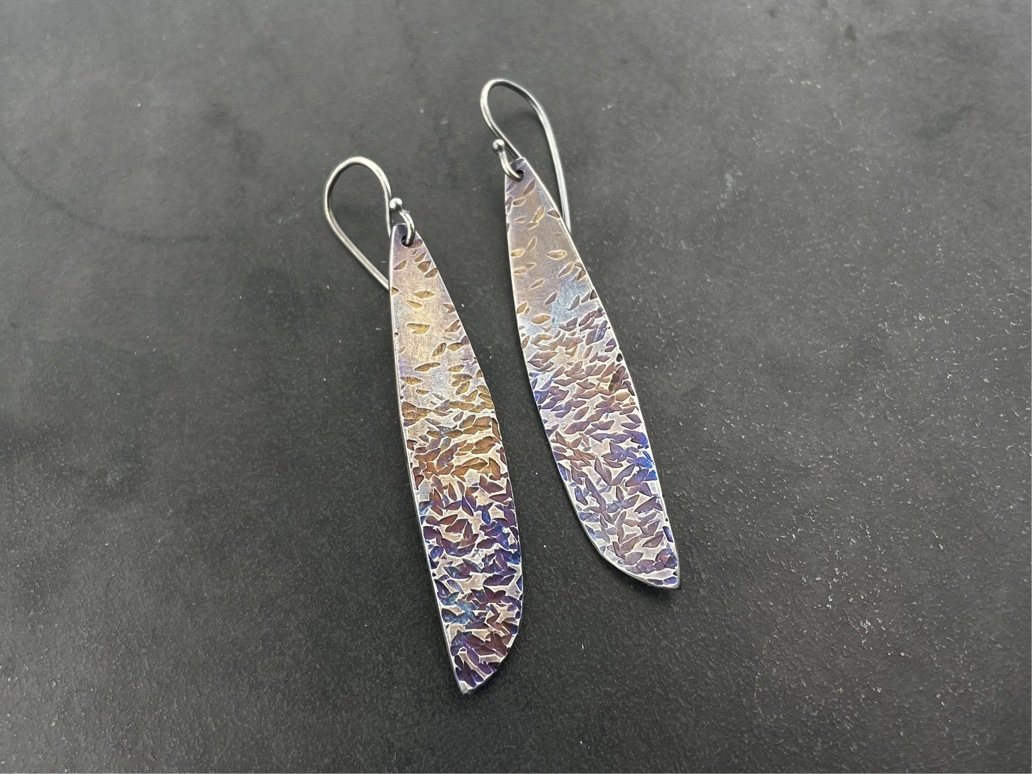 Pair of long, thin earrings with textured surface on a dark gray background